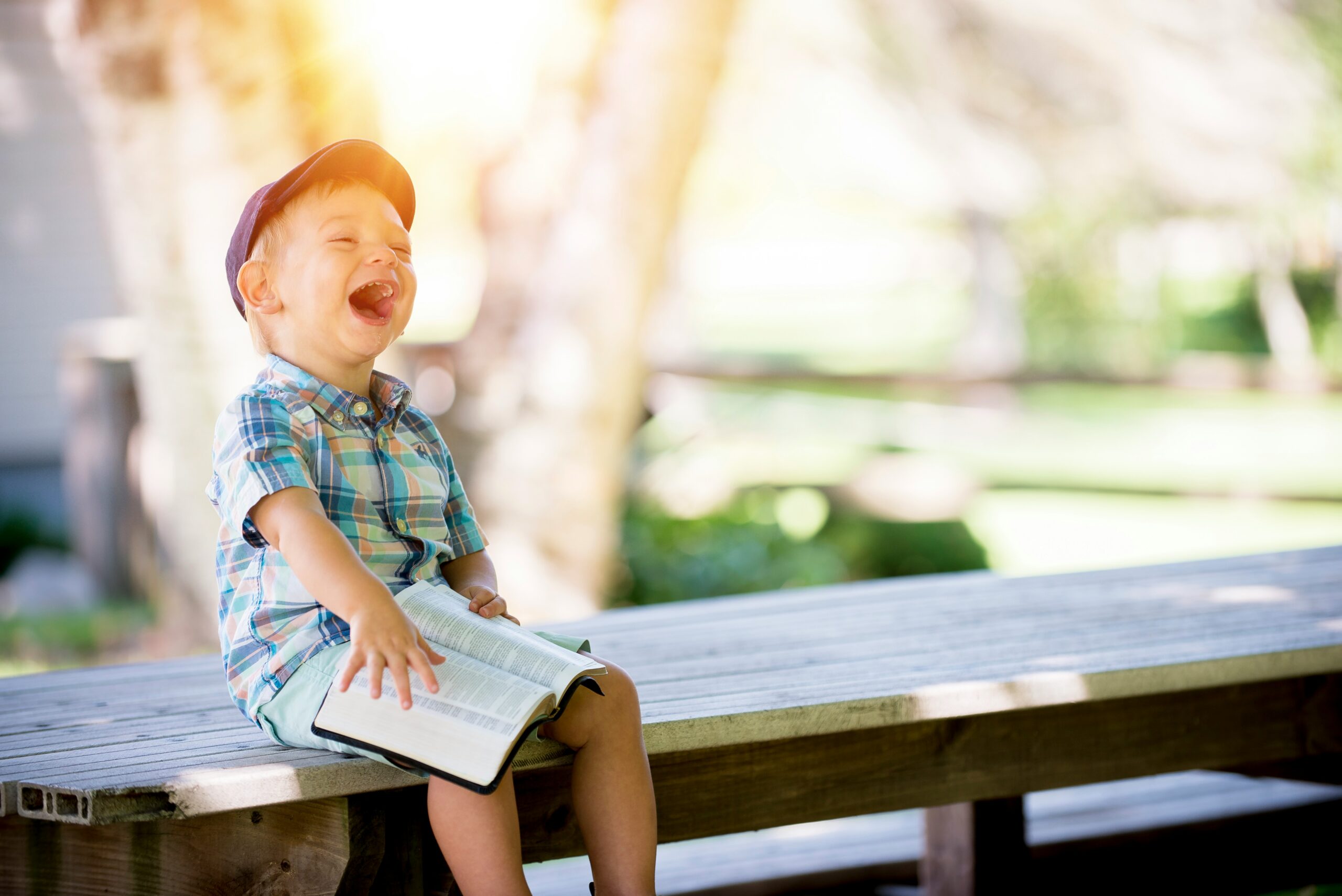A small child sitting on a bench with a book outdoors, laughing